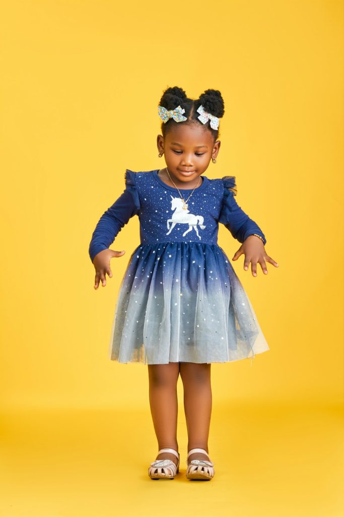 A little girl stands gracefully in a blue gradient dress with star details, captured against a bold yellow background.